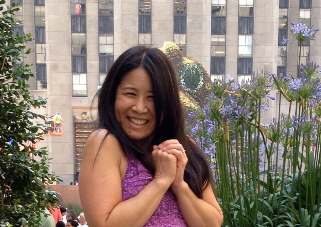 Image of Susan Wong of innerVelocity. She is an Asian woman with long dark hair wearing a pink sleeveless. She is smiling with her hands together in front of a tan building with many windows.