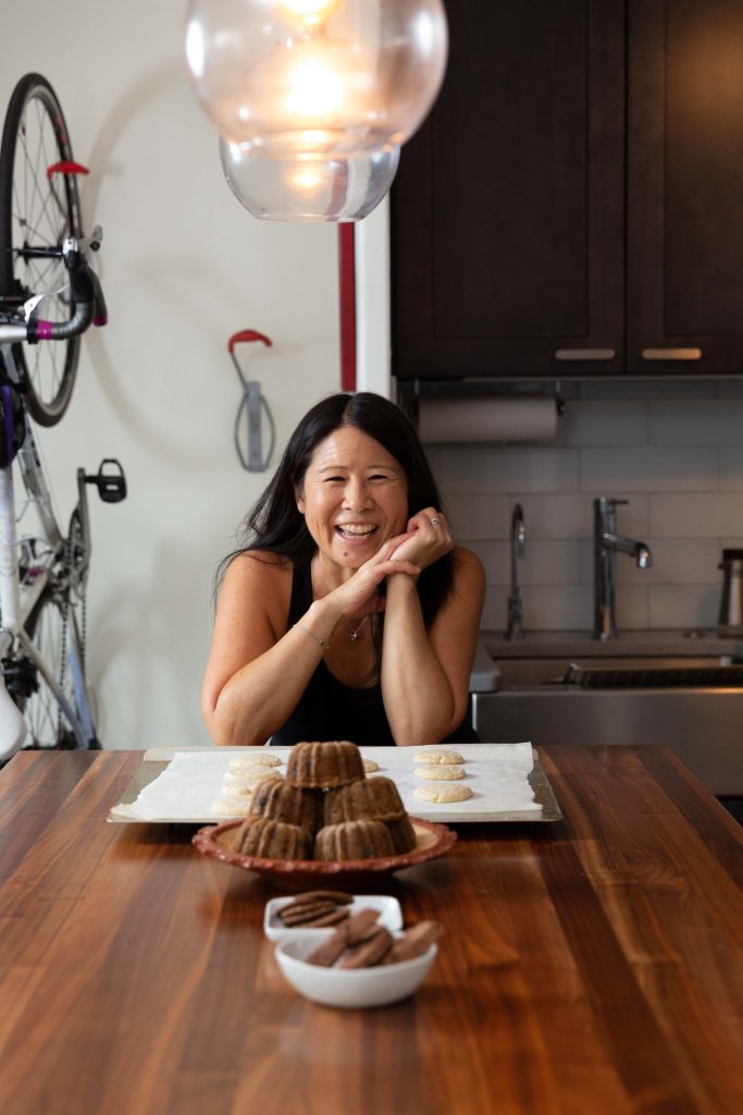 Susan Wong, who has long, dark hair and is smiling wearing a tank top. She is sitting at a wooden table in front of a silver baking dish with cookies on white parchment paper. There are also mini Bundt cakes piled on a plate.
