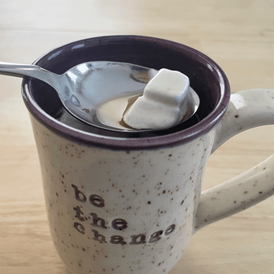 Image of a white speckled mug of hot chocolate with a silver teaspoon that contains two white mini marshmellows for innerVelocity.bike
