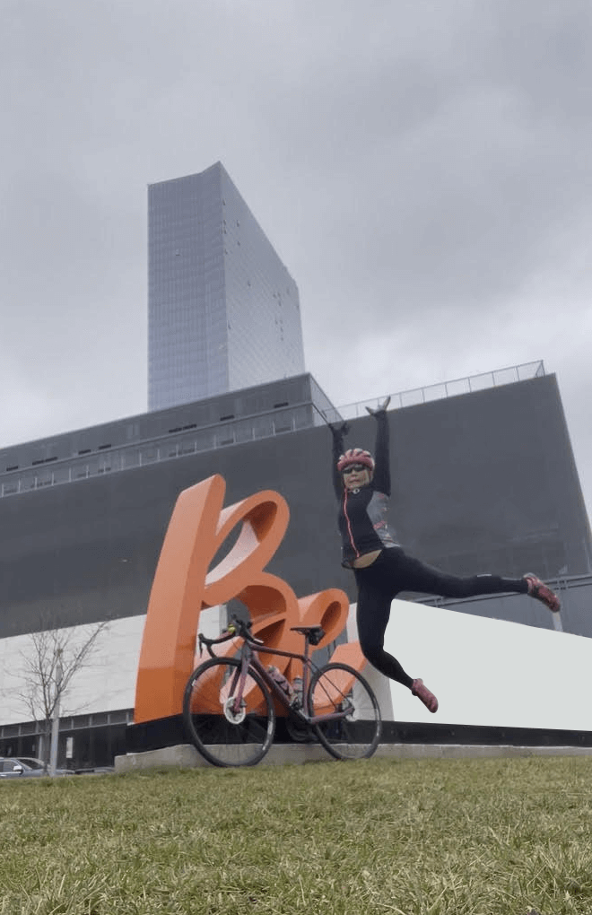 Image of Susan Wong of innerVelocity jumping in the air in front of a two wheeled bicycle that is leaning against a sculpture of the words Be.
