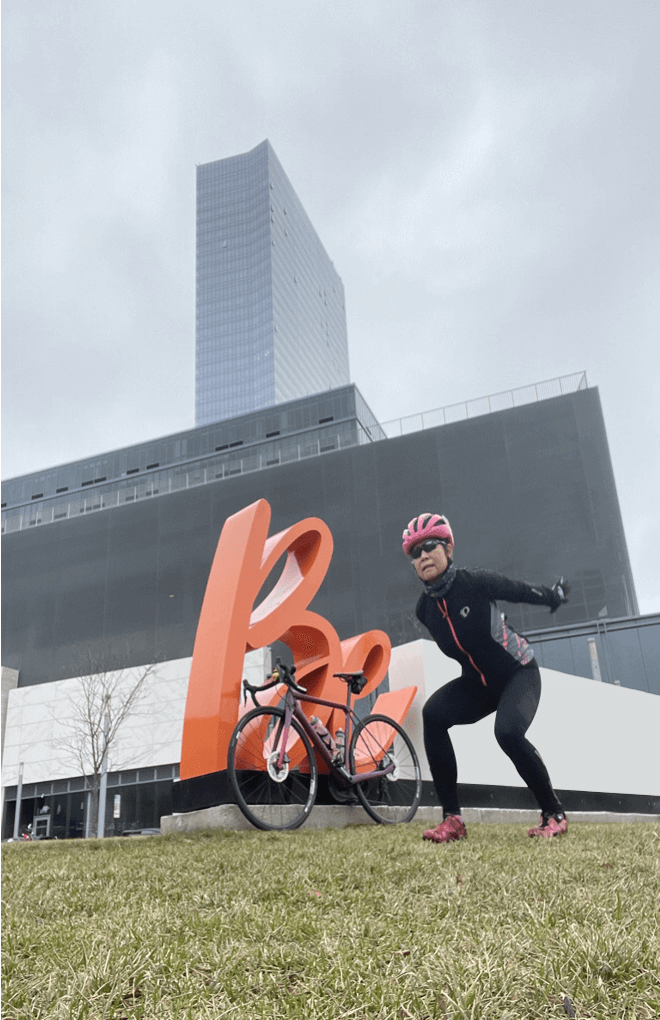 Image of Susan Wong of innerVelocity squatting in front of a two wheeled bicycle that is leaning against a sculpture of the words Be.